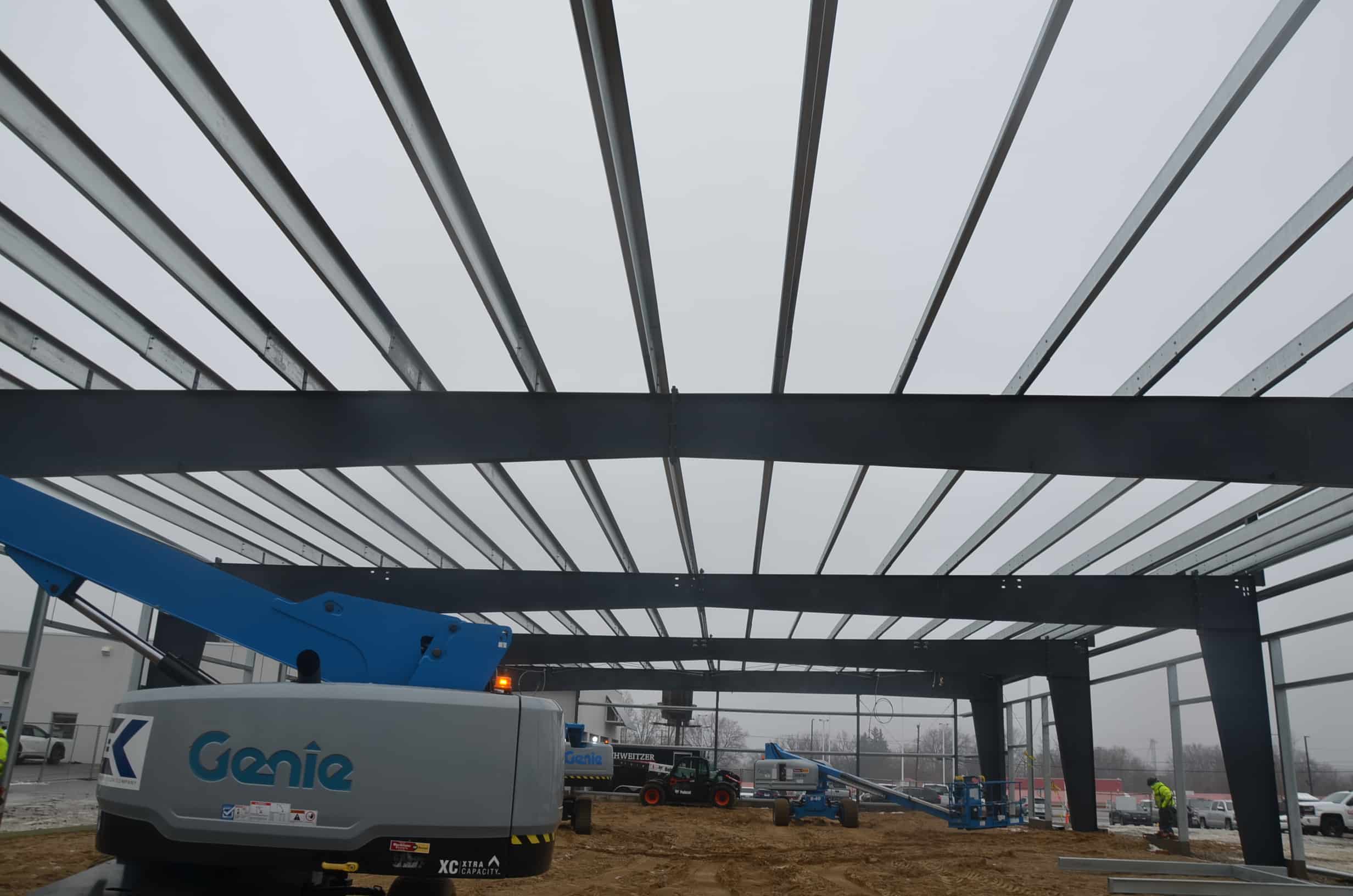 Steel beams form the frame of a building under construction, with heavy equipment and workers visible on a cloudy day at the job site.