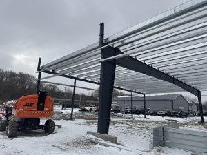 A steel building frame under construction on a snowy site, with an orange JLG lift nearby and several cars parked in the background.