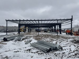 Steel framework of a building under construction on a snowy lot, with steel beams and construction equipment in the foreground.