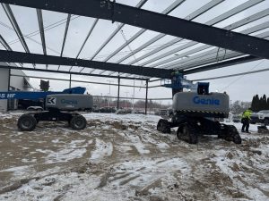 Two Genie boom lifts on a snowy construction site under a partially built metal structure, with workers standing nearby.