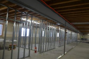 A large room under construction with exposed wooden ceiling beams and metal wall framing; construction materials and a fire extinguisher are visible.