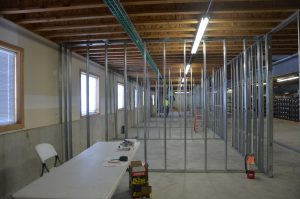 Room under construction with exposed ceiling beams, metal wall framing, some tools and materials on a folding table, and a ladder in the background.