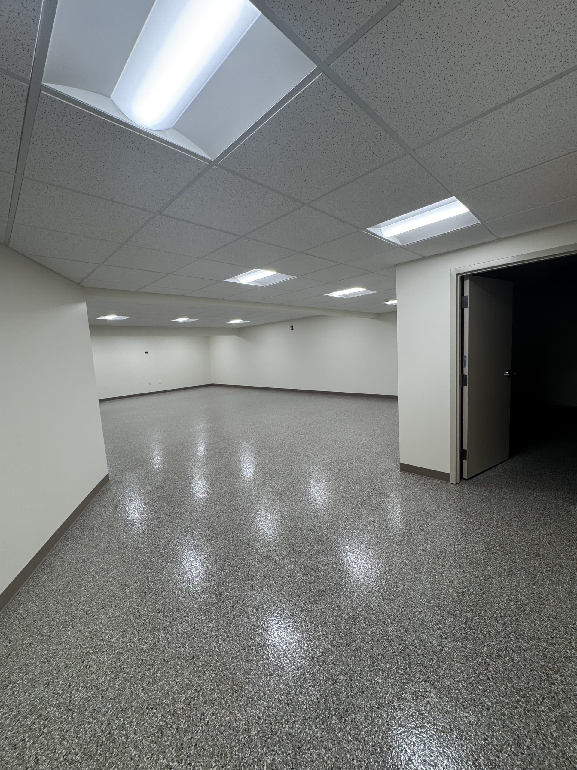 Empty room with speckled flooring, beige walls, fluorescent ceiling lights, and an open door leading to a darkened adjacent space.