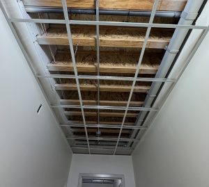 Exposed ceiling framing with a grid for a drop ceiling, showing pipes, wires, and wooden beams in a small room with white walls.