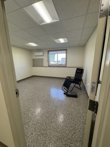 A nearly empty room with a recliner chair, a window, a closed door, and speckled flooring under fluorescent ceiling lights.