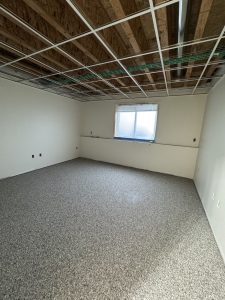 Unfinished room with exposed ceiling grid, bare walls, carpeted floor, and a single window letting in daylight.