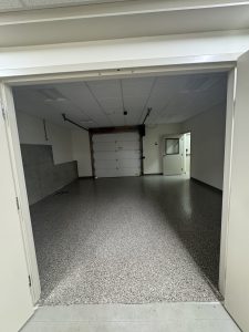 Empty garage with speckled flooring, white walls, a closed white garage door, overhead lights, and a small window near a doorway to another room.