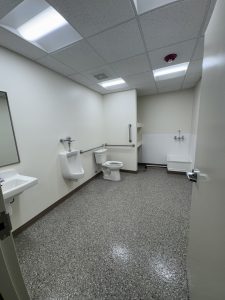 Accessible men’s restroom with two urinals, one toilet with grab bars, a sink, bright ceiling lights, and speckled floor.