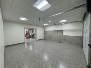 Empty garage with speckled floor, white ceiling tiles, fluorescent lights, and an open doorway leading to a storage area with various items visible in the background.