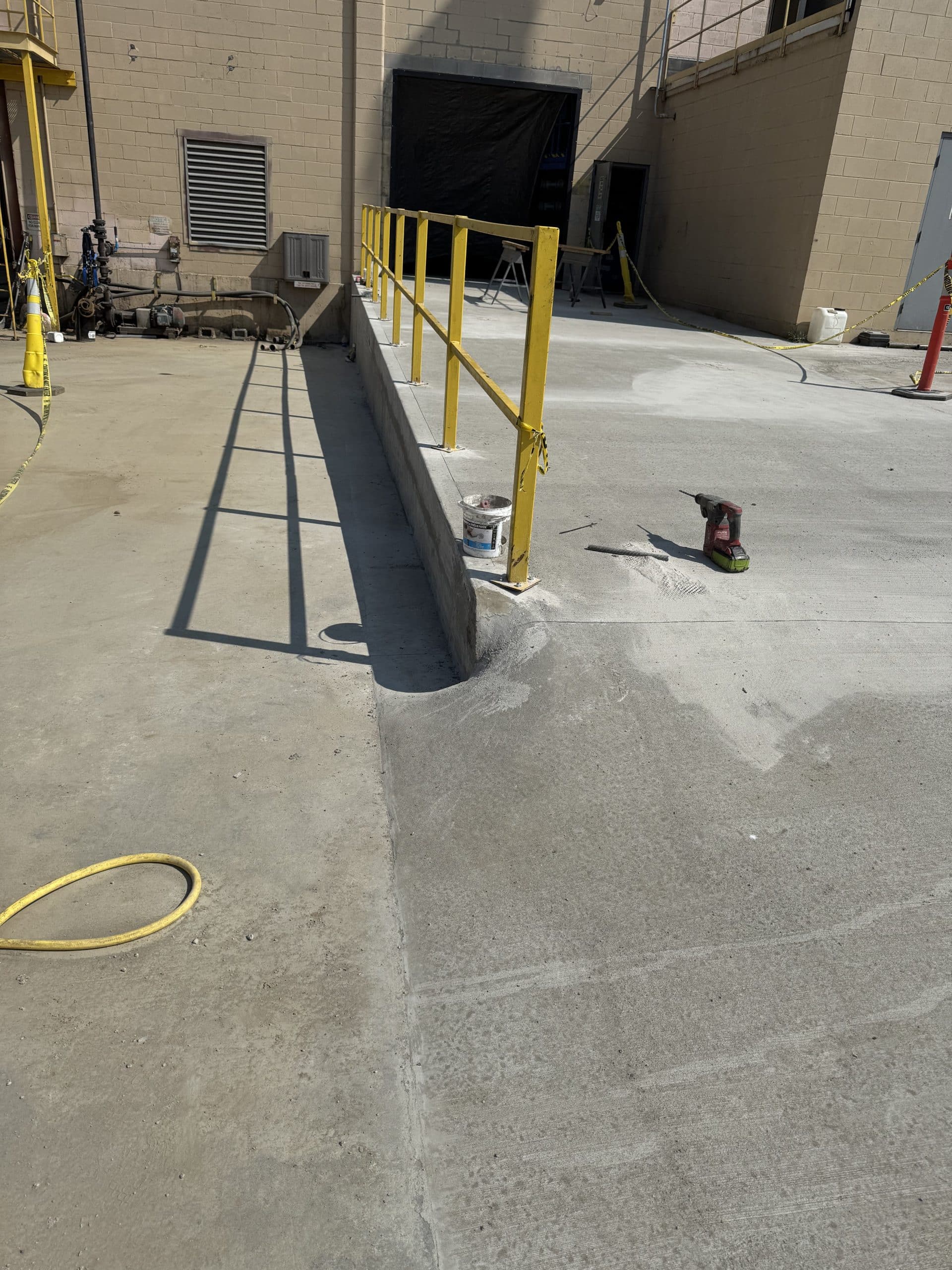 Concrete ramp with yellow handrails leads to a building entrance. A can, brush, and power tool sit on the ground. Utility pipes and cords are visible along the wall.