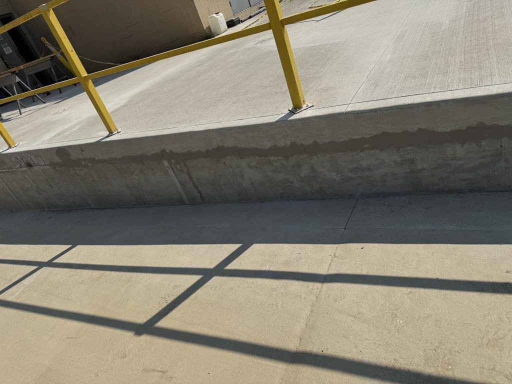 Concrete ramp beside a yellow metal railing, with shadows cast on the ground. A building and some equipment are visible in the background.